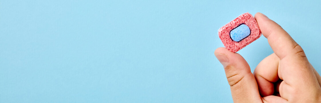 A close-up of a hand holding a single red and blue dishwasher detergent tablet against a light blue background. Wide panoramic banner format with generous copy space on the left