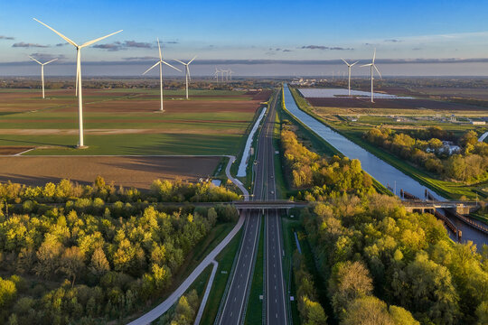 Aerial view of a highway stretching through rural fields with wind turbines, a canal, and lush green trees under a clear sky in Groningen, Netherlands.