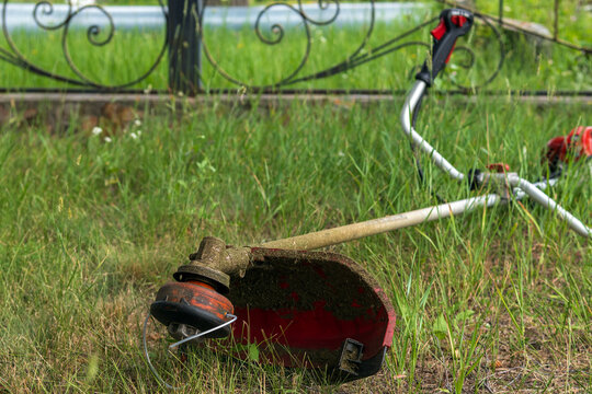 Gas string trimmer lying on a green lawn by a metal fence. Close-up of a dirty petrol grass trimmer or brushcutter resting on uncut green grass near a decorative wrought-iron fence. 