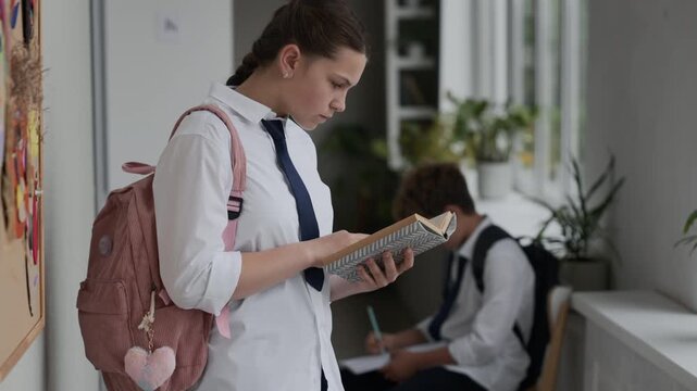 Caucasian teenage girl wearing school uniform standing in hallway and reading book while classmate studies in background. Perfect for educational marketing and school promos