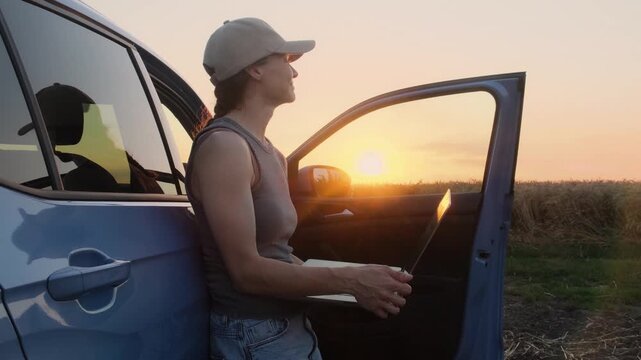A young farmer woman in work clothes stands with a laptop next to the car in the fields at sunset. She is tired, but satisfied with the result of her work.