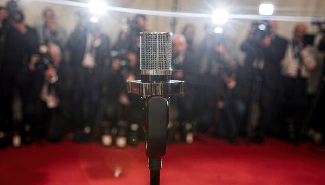 Crisp closeup on a sleek podium microphone during a red carpet media day with outoffocus photographers and flashing cameras behind.