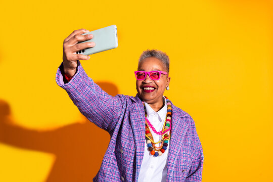 Senior woman taking selfie with smartphone on colorful background - Old hispanic grandmother wearing fashionable modern clothing on colorful background, concepts about elderly, aging, diversity 