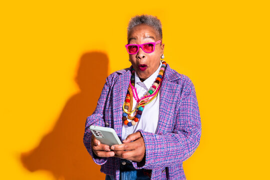 Senior woman reacting with surprise using smartphone - Old hispanic grandmother wearing fashionable modern clothing on colorful background, concepts about elderly, aging, diversity 