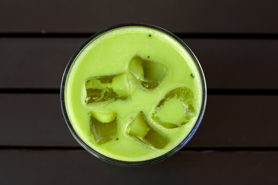 Top view of iced matcha latte in short glass with five large ice cubes submerged in pale creamy green liquid, placed on dark wooden slatted table