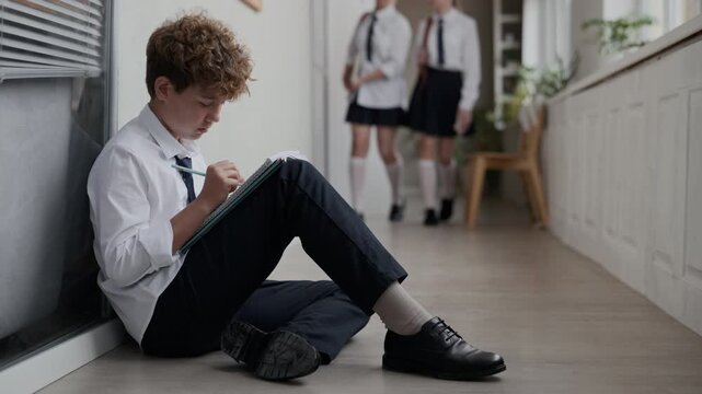 Caucasian teenage boy sitting on floor in school hallway and writing in notebook with classmates walking past. Ideal for educational campaigns or student mental health concepts