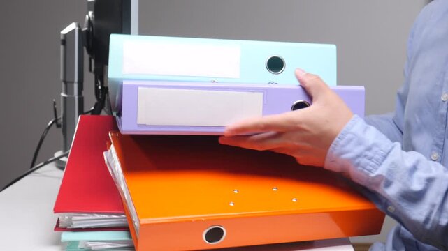Employee hands bringing a stack of colorful ring binders and document folders to an office desk, illustrating administrative work, data management, and the burden of paperwork