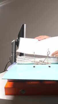 Vertical video person's hands organizing important business documents, inserting paper sheets into a ring binder for efficient archiving and office management, ensuring proper organization