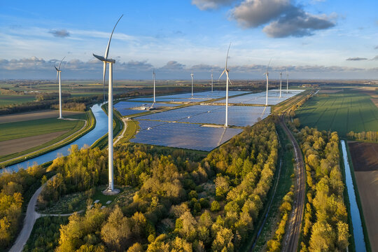 Aerial view of wind turbines and a large solar panel array situated in a rural landscape with green fields, a canal, and trees in Groningen, Netherlands.