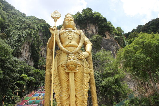 Giant golden statue of deity standing in front of limestone cave entrance stairs.
