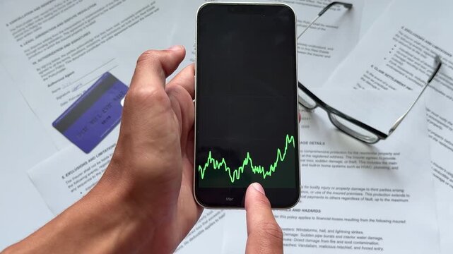 Hand holds smartphone displaying an upward-trending green line graph, symbolizing optimal equity growth tracking amidst financial documents and a credit card.