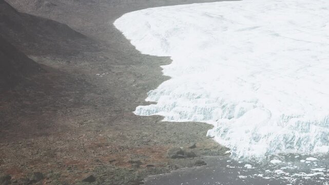 A large glacier extends towards the barren ground in a secluded Arctic region. The ice contrasts with the rocky terrain. It is summertime, and the landscape shows signs of melting ice.