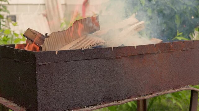 Bright orange flames and thick white smoke rise from dry firewood burning inside an old rusty metal barbecue grill. Cardboard and paper are used for quick ignition of logs in the backyard.