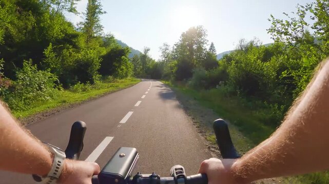 POV of cyclist riding on asphalt bike path at sunset in summer. Slovenia. First person view of male hands on bicycle handlebar cycling on road in green fields, forest. Active sport outdoor lifestyle