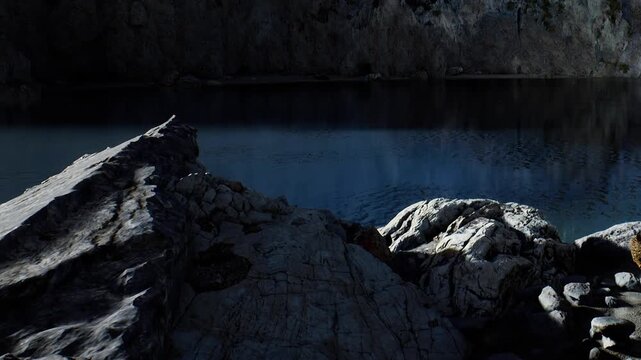 The scene shows a rocky shoreline with dark stones and calm water reflecting the surroundings. It is dusk and shadows linger over the natural features of the area.