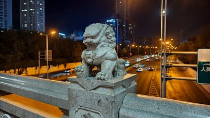 A traditional stone guardian lion (Fu Lion) sculpture standing on a bridge railing in the city center of Urumqi, Xinjiang, China. In the background, a busy highway with car light trail © Alexandra