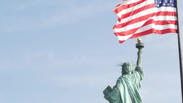 Statue of Liberty on Liberty Island, New York, United States. Cooper monument from behind, democracy and patriotism symbol near Manhattan, NYC USA. American flag waving in wind. Independence concept.