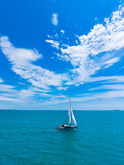 Fototapeta premium Sailboat moves through the blue water under a bright sky with white clouds in the daytime near the coast