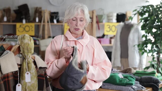 Mature woman choosing a warm fur hat in a clothing store. Elderly woman buys warm clothes for the winter. High quality 4k footage
