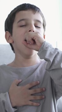 Child sitting on sofa at home and holding hand on chest. Male having asthma attack, difficulties with breathe, feeling severe pain or dyspnea