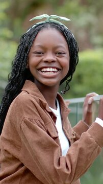 Vertical video: Smiling girl leaning on metal rail in park, camera framing and showing brown jacket