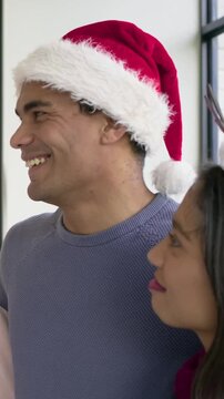 Vertical video: Hearing offscreen voice, pair smiling, laughing near window with Santa hat, antlers