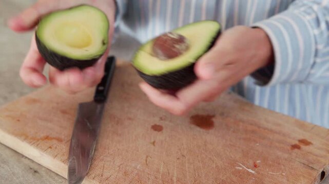 Close up of woman hands opening it in half  and cutting fresh avocado with knife on wooden chopping board in kitchen, preparing healthy organic food recipe for salad at home