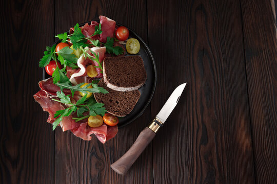 Sliced jamon and bacon on a dark plate with a pocket knife with wooden handle, rustic background.