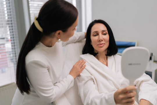 Friendly beautician doctor providing personalized consultation to female client in specialized treatment chair, reviewing her skin appearance in mirror inside contemporary beauty salon.