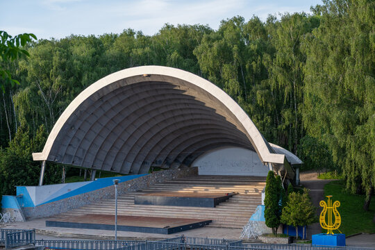 A large, arched concrete bandshell known as Spivoche Pole stands empty, surrounded by lush green foliage in a tranquil public park setting.