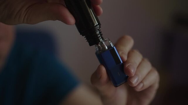 Close-up view of man hands pouring e liquid from bottle into tank of modern blue vape pod system, preparing device for use and demonstrating vaping maintenance. Shooting in slow motion
