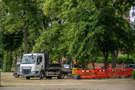 Builders&rsquo; truck with a tipper body parked on a grass area beside a construction site. The area is cordoned off with red plastic safety barriers, while a small yellow excavator operates in the back