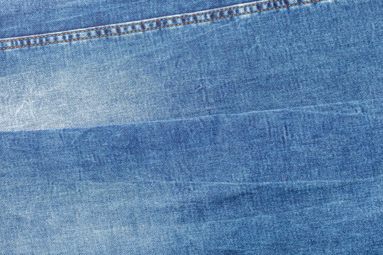 High-angle view of bright blue denim fabric layers. Features an explicit horizontal double stitched seam across the top section, fine diagonal twill weave, and soft gradient fading.