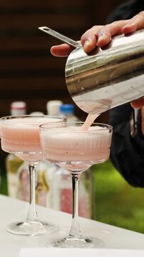 Vertical video. Bartender pours pink cocktail from shaker into glass at outdoor wedding bar.