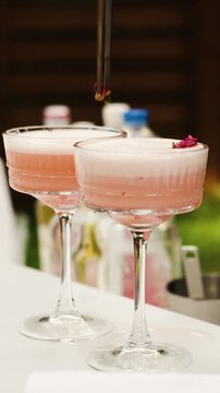 Vertical video. Bartender decorates pink cocktail with flower petal using tweezers at outdoor bar.