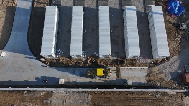 Five white shipping containers neatly arranged at a construction site with heavy machinery and prepared ground