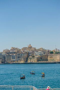 Traditional Maltese dghajsa boats crossing the Grand Harbour with historic limestone fortified town skyline in Malta on a clear summer day