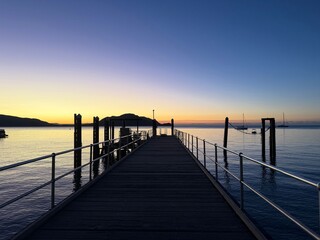 Obraz premium Wooden pier jetty leading to calm ocean at sunset with sailboats and mountain silhouette against colorful sky