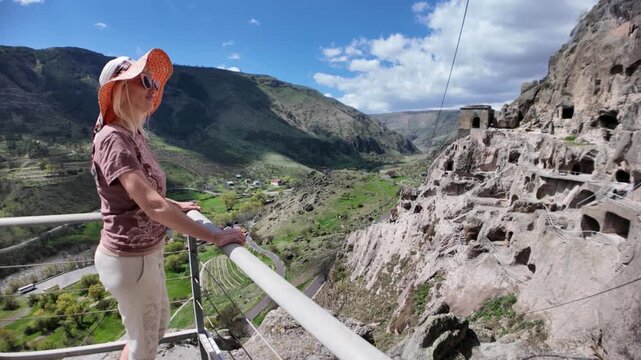Woman standing on a viewing platform, admiring the ancient multi-story Vardzia cave monastery carved into the rocky hillside and the scenic green valley of Erusheti Mountain in Georgia