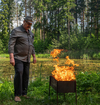 Man outdoors tending a grill with bright flames rising from it while standing in a forest area near a pond surrounded by dense greenery