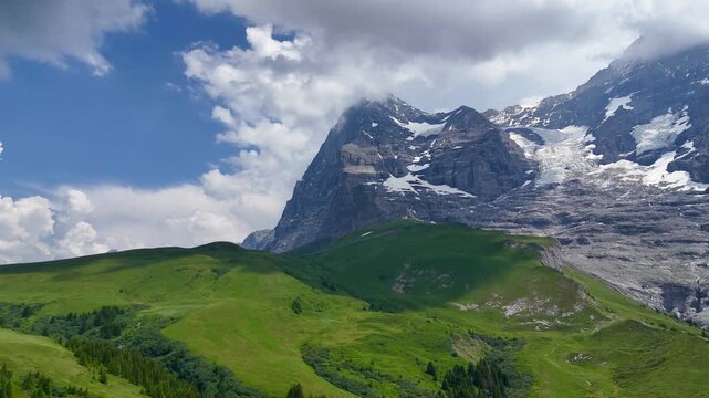 A breathtaking panoramic view of the iconic Eiger mountain face towering over lush green hills and alpine pastures in the Swiss Alps under a blue sky with dynamic clouds during a beautiful summer day.