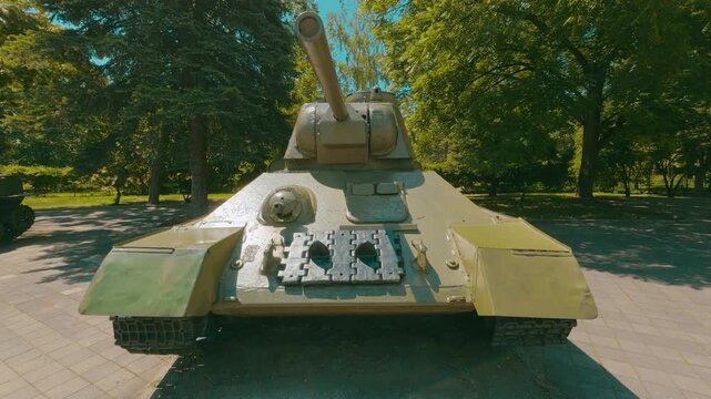 A frontal view of a battered T-34 tank parked on paved ground surrounded by lush green trees. Sunlight casts harsh shadows. The olive-green paintwork shows visible wear