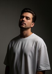 Obraz premium A dramatic studio portrait of a man with a mustache looking down at the camera, wearing a loose white t-shirt against a dark grey background with moody lighting