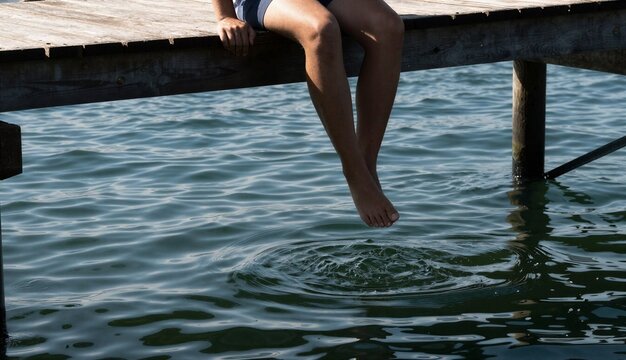 Person&rsquo;s bare legs and feet dangling from a weathered wooden dock over water
