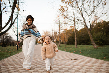 Fototapeta premium Funny child girl 2-3 year old run away from her mother in autumn park wearing knitted clothes outdoors. Motherhood. Fall season.