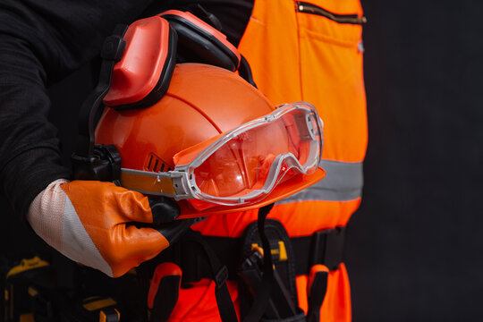 Construction worker holding personal protective equipment including safety helmet, goggles and earmuffs on dark background. Occupational safety, PPE compliance, protection and industrial construction.