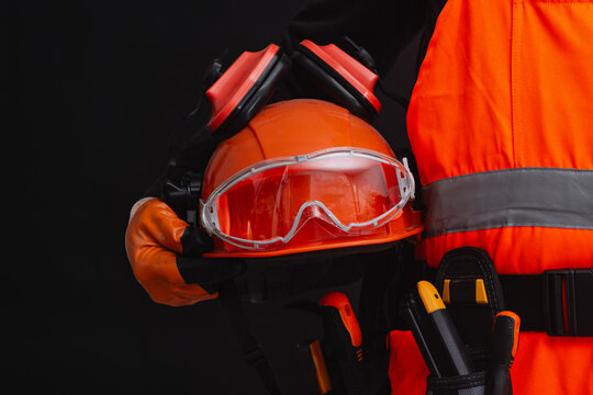 Construction worker holding personal protective equipment including safety helmet, goggles and earmuffs on dark background. Occupational safety, PPE compliance, protection and industrial construction.