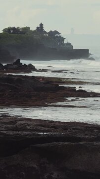 Misty Coastal Tanah Lot Temple Cliff At Sunrise, Solitary Pilgrim Imagined On Rocks, Waves Crashing Over Dark Basalt, Distant Hindu Shrine Silhouette, Tranquil Meditative Atmosphere, Vertical video
