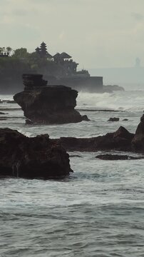Rocky Offshore Stacks Under Overcast Sky, Tanah Lot Temple Silhouette In Background, Turbulent Sea Rolling Over Jagged Basalt Reefs, Cinematic Moody Palette, Bali island, Indonesia, vertical video