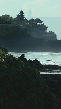 Rocky Offshore Stacks Under Overcast Sky, Tanah Lot Temple Silhouette In Background, Turbulent Sea Rolling Over Jagged Basalt Reefs, Cinematic Moody Palette, Bali island, Indonesia, vertical video